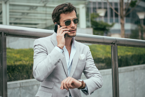 Young businessman in a formal jacket, waiting for a friend at the city square, while talking on a mobile phone.