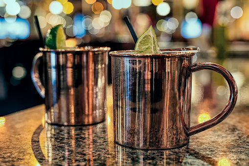 Two Moscow Mule cocktails in copper mugs on a marble table in a bar. The drinks are garnished with black straws and a lime.