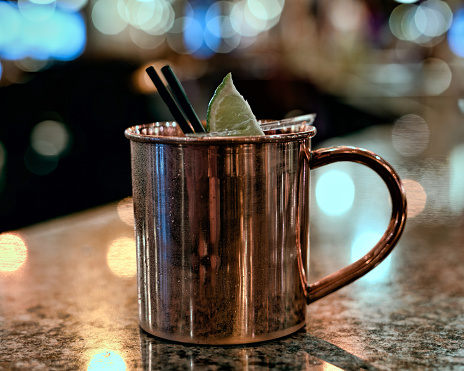 A Moscow Mule cocktail in a copper mug on a marble table in a bar. The drink is garnished with black straws and a lime.