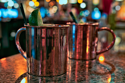 Two Moscow Mule cocktails in copper mugs on a marble table in a bar. The drinks are garnished with black straws and a lime.