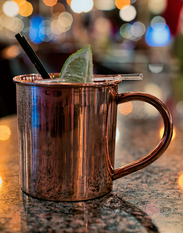 A Moscow Mule cocktail in a copper mug on a marble table in a bar. The drink is garnished with black straws and a lime.