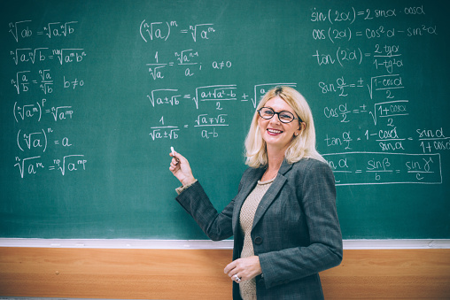 Young happy smiling woman school professional educator, university mentor, instructor or teacher, female coach standing at desk in front of chalkboard in modern college classroom or work office.