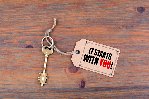 Key and a note on a wooden table with text - It Starts With You