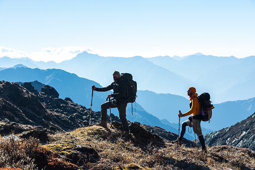 Silhouettes of two Hikers staying on rocky and grassy Ridge with Backpacks and other Gear expressing Energy and Happiness. Layered Mountain Valley View on Background