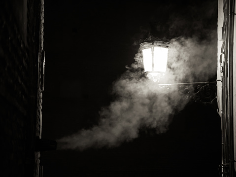 The smoke from a fireplace merges with the light of a street lamp during the night.