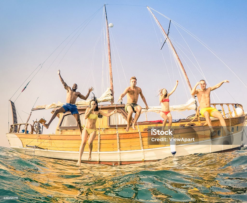 Young multiracial happy friends jumping from sailing party boat Frontal view of young multiracial friends jumping from wooden sailing boat on sea trip - Rich happy guys and girls having fun in summer party day - Exclusive vacation concept - Warm vintage filter Friendship Stock Photo
