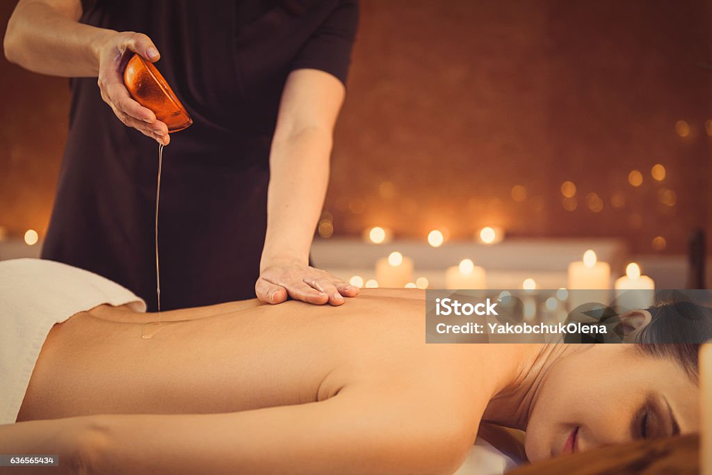 Calm girl getting treatment at wellness center Happy young woman is enjoying massage at spa. She is lying and smiling. Beautician is standing and pouring oil from bowl Massaging Stock Photo