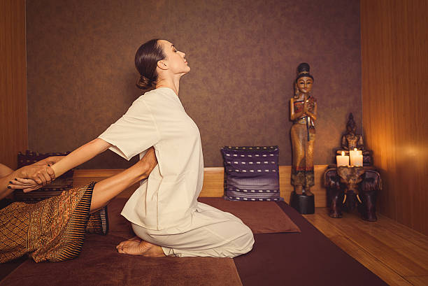 Serene girl enjoying thai massage Professional thai masseuse is stretching female body. She is raising leg to her back. Young woman is sitting on floor. Her eyes are closed with pleasure thai ethnicity stock pictures, royalty-free photos & images