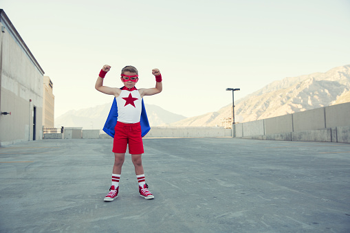 A young boy wearing a red superhero cape and mask stands on a parking garage in Utah; as he flexes his muscles, he imagines taking off to help make a brighter future. He has a star on his chest and is showing strength against adversity.