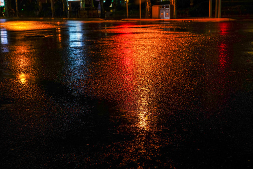 Rainy night in the big city, light from the shop windows reflected on the road on which cars travel. View from the level of asphalt