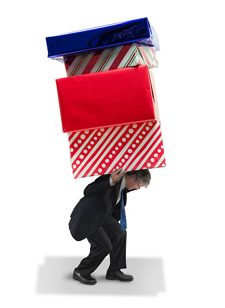 Man carrying giant gifts Christmas and birthday financial spending burden Man struggling to carrying stack of giant gifts representing financial burden of Christmas, Hanukkah, birthdays, anniversaries, parties, weddings and the constant spending that goes with them. hanukkah shopping stock pictures, royalty-free photos & images