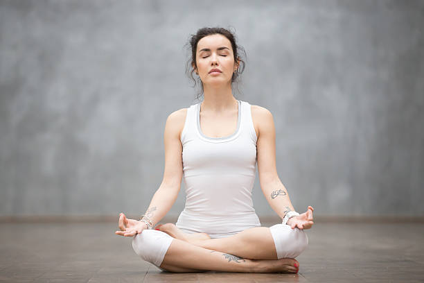 Beautiful Yoga: Half Lotus pose Front view portrait of beautiful young woman with floral tattoos working out against grey wall, resting after doing yoga exercises, sitting in ardha Padmasana, Lotus pose, relaxing. Full length sukhasana stock pictures, royalty-free photos & images