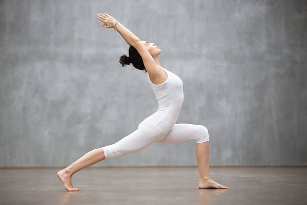 Beautiful Yoga: Warrior one pose Side view portrait of beautiful young woman wearing white tank top working out against grey wall, doing yoga or pilates exercise. Standing in Warrior one pose, Virabhadrasana. Full length warrior position stock pictures, royalty-free photos & images