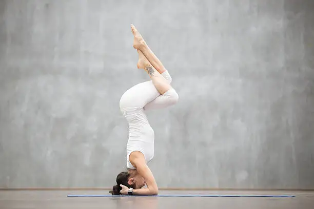 Portrait of beautiful young woman wearing white sportswear working out against grey wall, doing yoga or pilates exercise. Headstand, Garuda salamba sirsasana. Full length photo