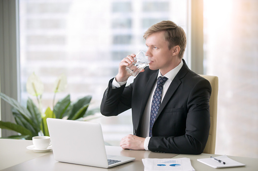Portrait of young handsome businessman working with laptop at the desk in the modern office, drinking water after a difficult talk, relaxing, keeping diet, plain water instead of coffee, thirsty