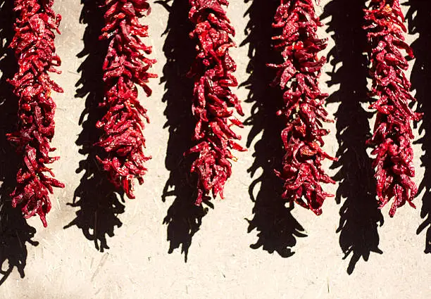 Red chili pepper ristras are hung to dry against a sunlit beige adobe wall in New Mexico. Copy space at the bottom of the frame.