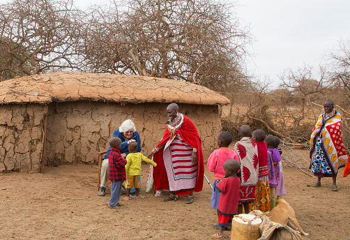 Selenkay. Kenya - October 9, 2016: Senior tourists in Maasai village. Greeting children. A wise haired European woman is sitting down and the children are coming up to be greeted the traditional way by touching their heads. Two women in traditional manga and jewelry super wise. In the background a typical hut made of coins dung.