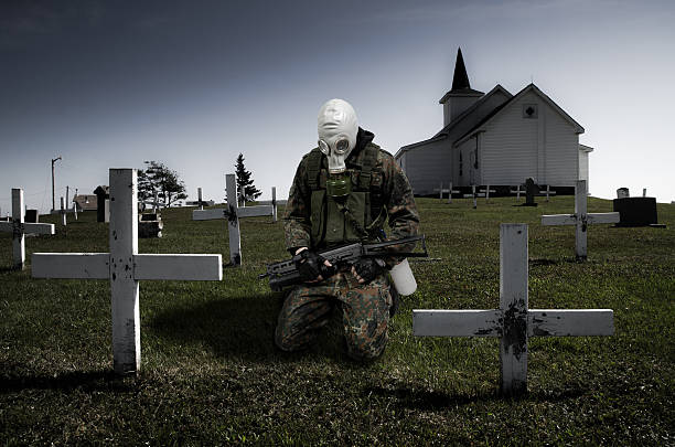 Soldier with gas mask kneeling on the grass in front of cross in cemetery during day of summer