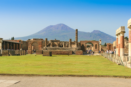 Pompeii, Italy - September 9, 2008: Ruins of Pompeii, Italy. Mount Vesuvius, green grass, trees, sightseeing tourists and vivid blue clear sky are in background. The Roman City of Pompeii was buried under a thick carpet of volcanic ash in the year 79 A.D when a Volcano Mount Vesuvius (Monte Vesuvio) has erupted. The eruption was one of the most catastrophic volcanic eruptions in European history. The volcano is located on the Gulf of Naples in Campania, about 5.6 mi east of Naples.