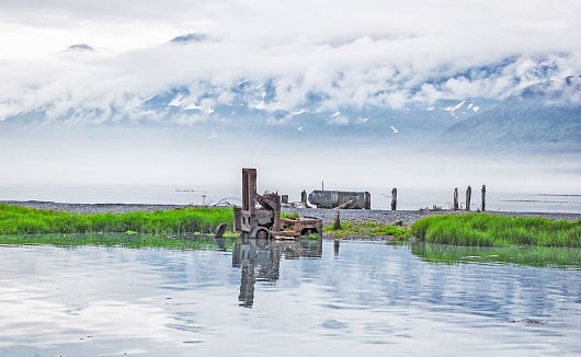 Picture of an abandoned forklift with reflection in the Prince William Sound, near Valdez, Alaska.  Fog can be seen rolling in at the base of the cloud covered mountain.