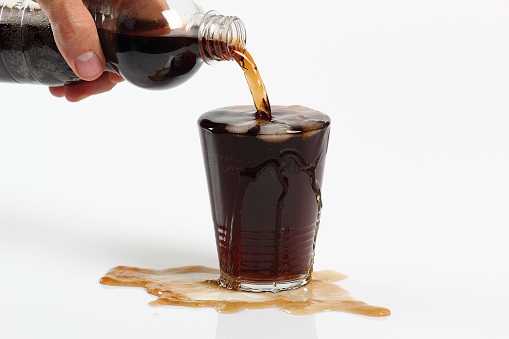 Human hand pouring cola from plastic bottle into overflowing glass with ice cubes. Isolated on white background.