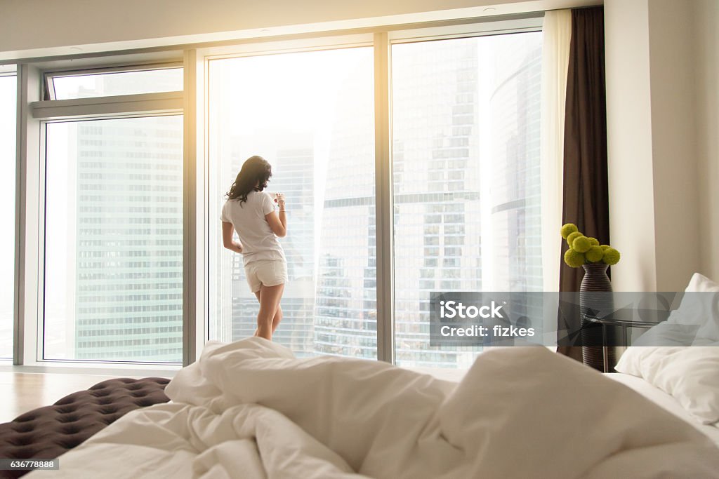 Young woman after wake up Unmade bed with white pillows and sheets, attractive casual girl, drinking coffee looking through window after waking up, making mornings enjoyable, new day is coming, good accommodation in big city City Stock Photo