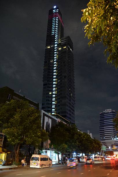 Bangkok, Thailand - October 6, 2016: Cars riding on a main street in the Silom district of Bangkok, Thailand. In the background, the tall and thin M Silom building, a luxury condominium 53-storey tower, height 198 mt. in the heart of the financial district. People sitting aside the street.
