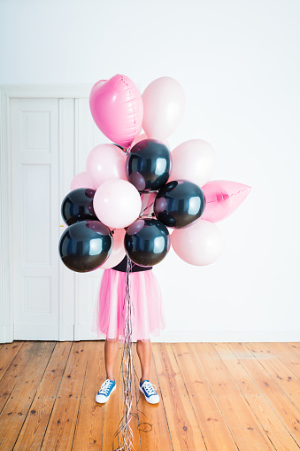 Young woman wearing black blouse and pink skirt jumping with bunch of pink and black balloons in front of her face.