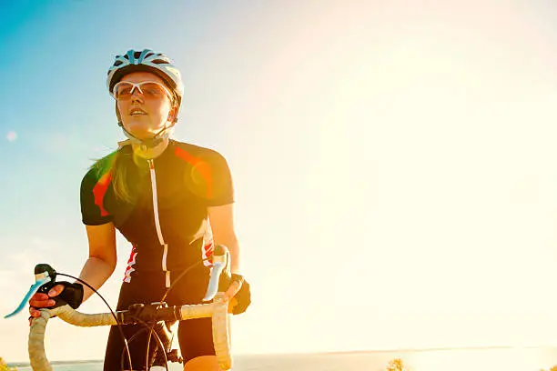 Close up portrait of female bike rider, who rides her bike up a hill. She wears helmet and sports glasses. She holds on to the handle bar. The sun sets in the background over the sea. Lots of copy space.