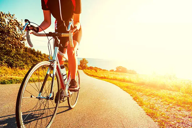 Close up of a athlete who rides her bike up a steep hill. The sun shines as her feet hits the pedals and she drives the bicycle forward. In the background is the sun setting over the sea.