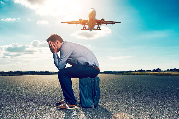 Sad passenger waits with suitcase for plane on runway Male passenger sits on his suitcase on an airfield runway and covers his head in his hands. He looks tired and frustrated. Perhaps his plane got cancelled or delayed. Perhaps he missed his plane. Sun shines in the background where an airplane takes off. cancellation photos stock pictures, royalty-free photos & images