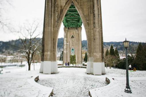 A fresh layer of snow fallen on the ground underneath the historic Saint Johns Bridge in Portland Oregon. The St. Johns Bridge is a steel suspension bridge that spans the Willamette River in Portland, Oregon, USA, between the St. Johns neighborhood and the northwest industrial area around Linnton. It towers above the river at 408 feet tall and was constructed in 1929.