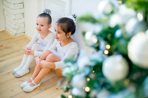 Two young ballet dancer sitting near Christmas tree on the wooden floor and smile