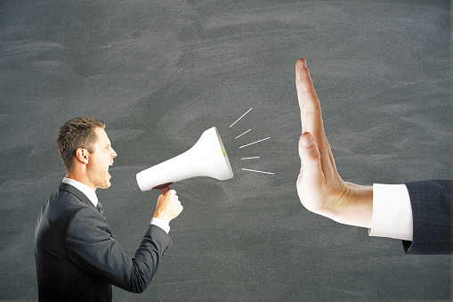 Irritated young businessman with megaphone screaming at hand showing stop gesture on chalkboard background. Protest concept