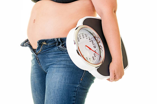 Fat overweight woman carrying a bathroom scale. Fat overweight woman wearing unzipped blue denim jeans carrying a bathroom scale under her arm side view close up on her stomach. fat zip stock pictures, royalty-free photos & images