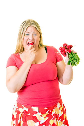 Overweight woman grimacing and eating healthy radishes on white.