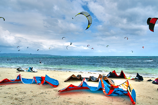 Colorful Kite surfing equipment laying  on a white sand beach in the Philippines with bright blue water and cloudy blue sky in the background. 
