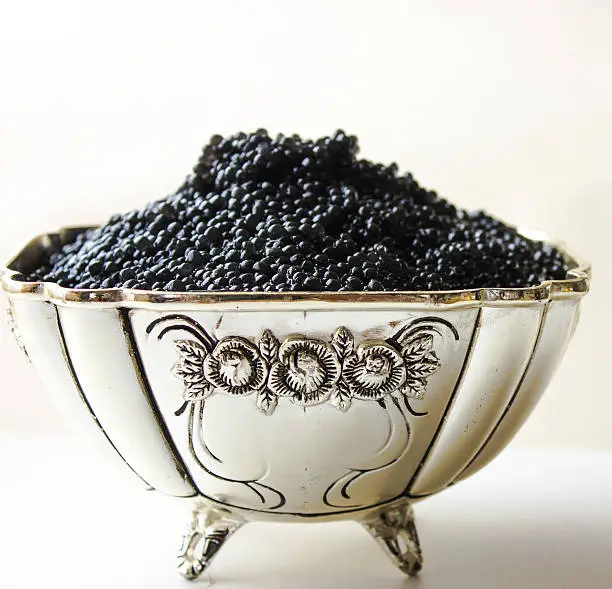 Black caviar in a silver bowl on a white background with a silver spoon and lemon