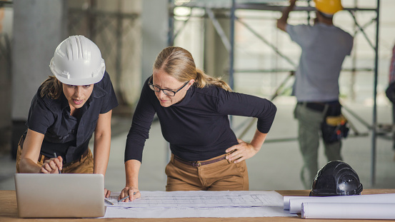 Front view of two women standing by the desk full of blueprints and laptop, construction worker in background.