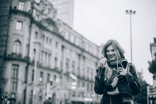 Beautiful young woman in a leater jacket and with a scarf standing on a town square on a cold day, listening to music on her mobile phone.