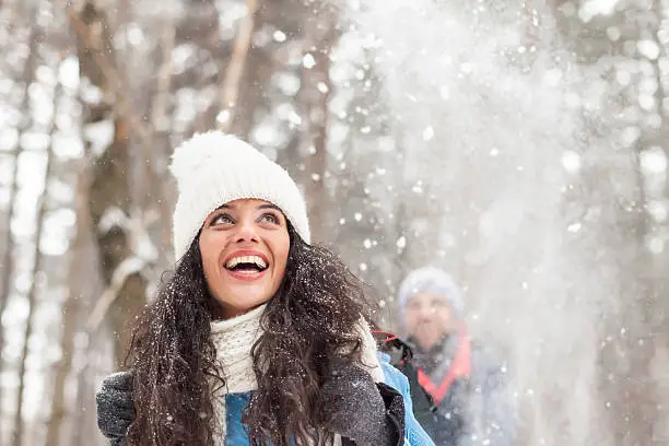 Happy young woman having fun in the snow forest. Man standing behind her. Both wear warm clothes. Snowflakes in the air. Snow trees on background.