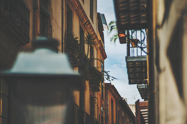 View of facades of residential houses on narrow street with sky behind with a lot of balconies, windows home plants, blurred lantern in front,  Barcelona historical district El Born, sunny summer day