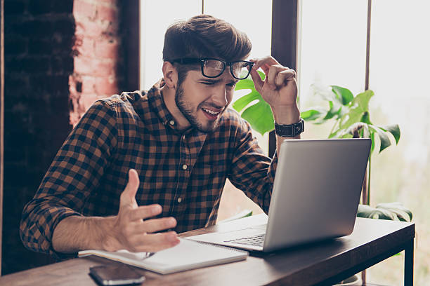 beau jeune homme stressé dans des lunettes à l’aide d’un ordinateur portable - manager anger table furious photos et images de collection