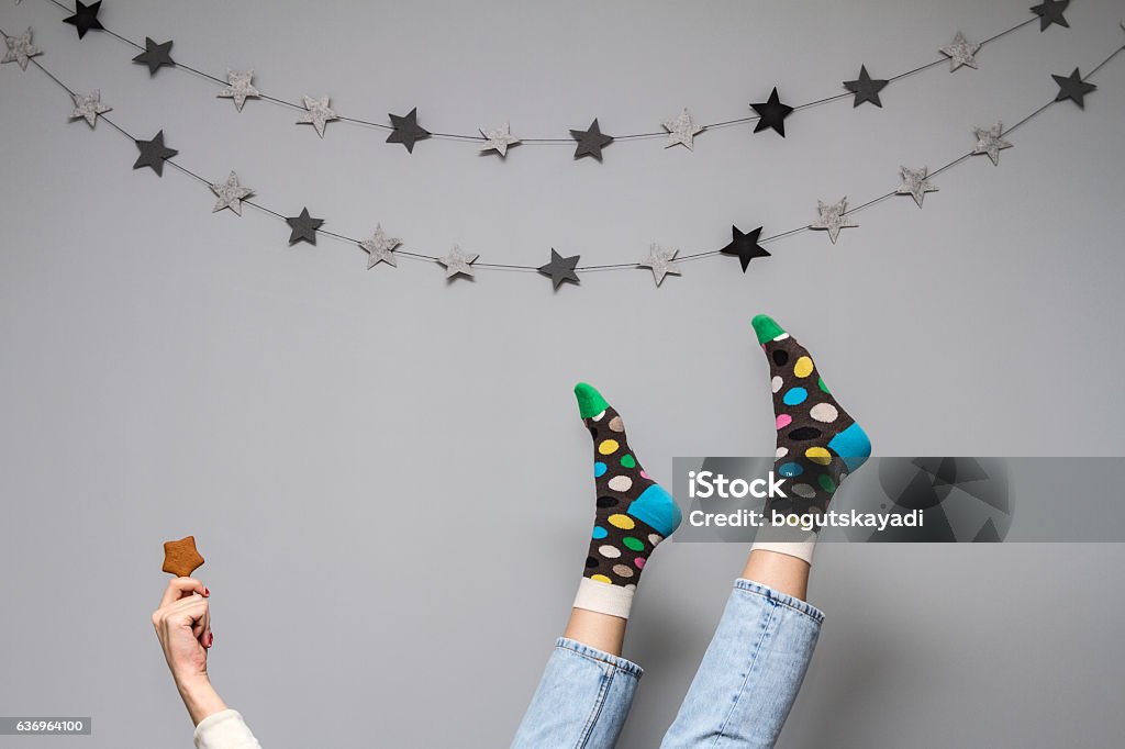 Women's legs in funny socks on a gray background. Women's legs in colorful funny socks and jeans on a gray background with the decor of the stars and ginger cookies in the shape of a star in her hand. Sock Stock Photo