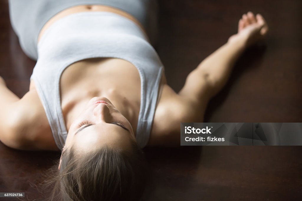 Shavasana yoga posture on the floor Beautiful young woman working out at home, doing yoga exercise on wooden floor, lying in Shavasana (Corpse or Dead Body Posture), resting after practice, meditating, breathing. Close up Breathing Exercise Stock Photo