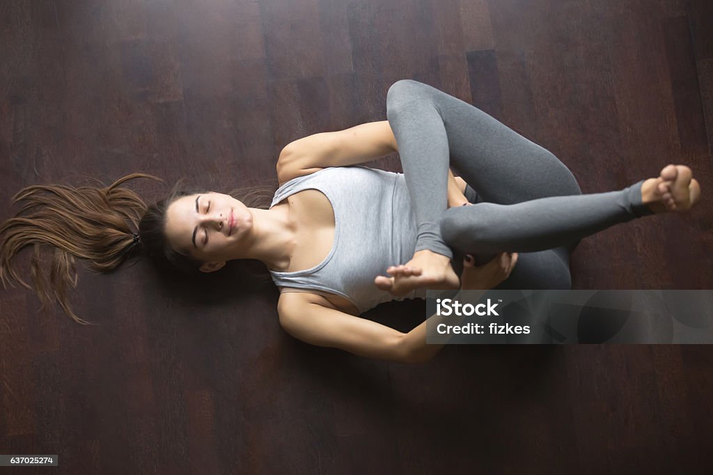 Top view of Eye of the Needle yoga pose Beautiful young model working out at home, doing yoga exercise on floor, lying in Eye of the Needle hip stretch pose (Dead or Reclining Pigeon posture), resting after practice. Full length, top view Stretching Stock Photo