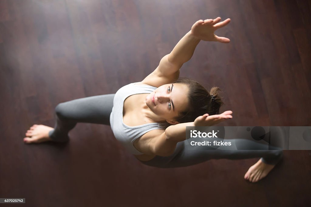 Top view of virabhadrasana 1 yoga Pose Beautiful young model working out in home interior, doing yoga exercise on wooden floor, standing in warrior I, virabhadrasana 1 pose. Top view. Full length Yoga Stock Photo