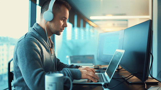 Closeup side view of a software developers working late at IT office. Mid 20's typical IT expert sitting at his desk and using multiple computers. He's listening to music on a headphones set.