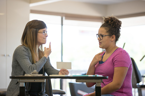 A young adult female speech therapist is helping a teenage patient inside of a clinic