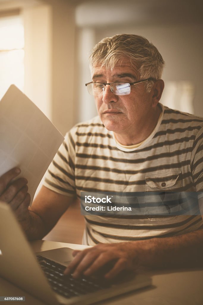 Senior man paying bills online on laptop. Elderly man working at home office in the late afternoon. 60-64 Years Stock Photo